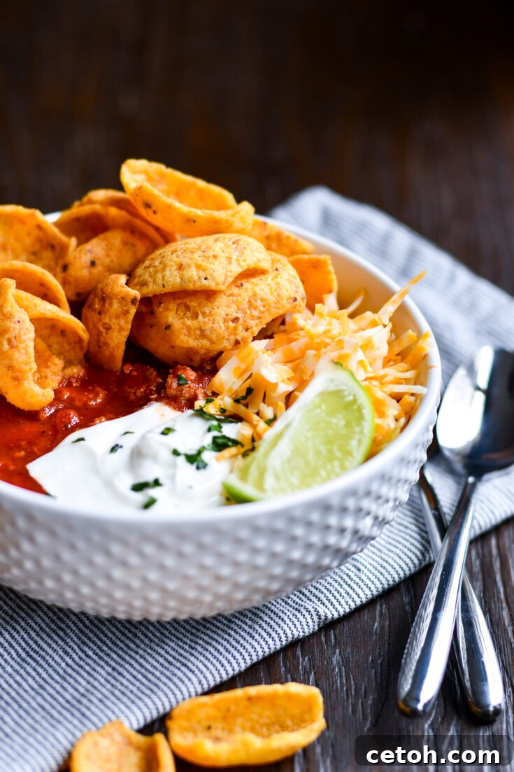 A vibrant bowl of Dad's Southern Homemade Chili, generously topped with shredded cheddar cheese, a dollop of sour cream, and a handful of crispy Fritos, is served on a rustic wooden surface, highlighting its hearty appeal.