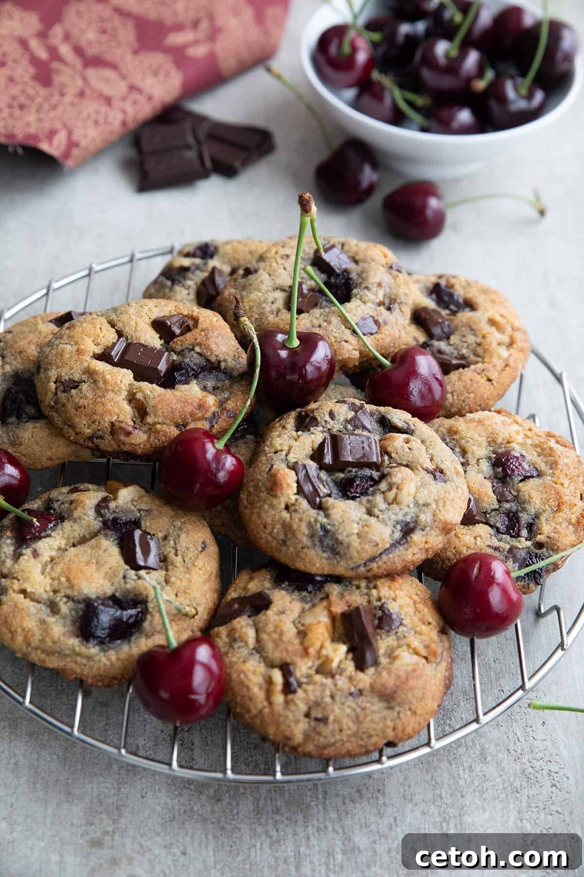 Decadent Keto Cherry Chocolate Chunk Cookies 2 Keto Cherry Chocolate Chunk Cookies on a cooling rack with a bowl of cherries in the background.