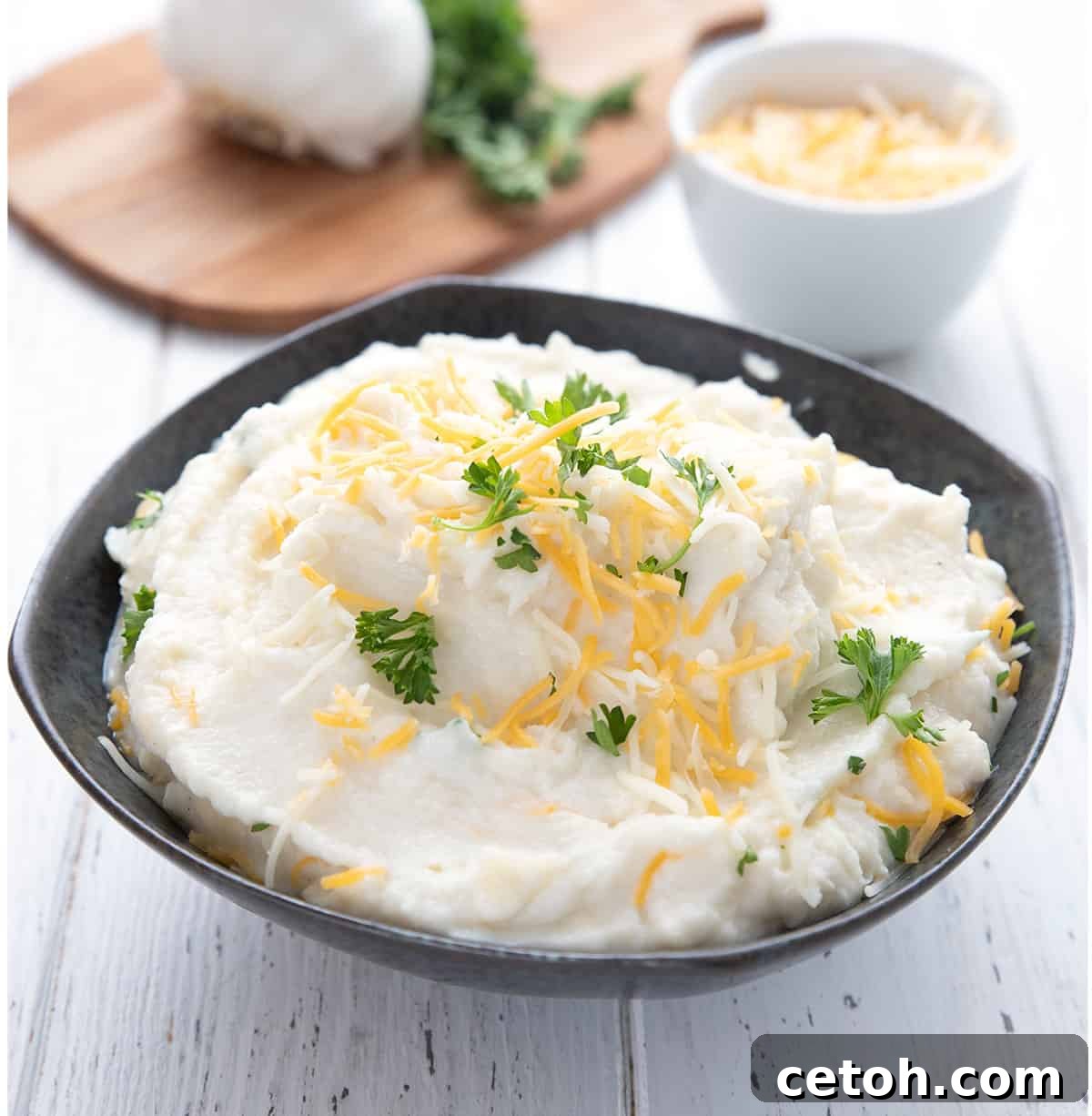 A black bowl filled with creamy mashed cauliflower topped with cheddar and parsley.