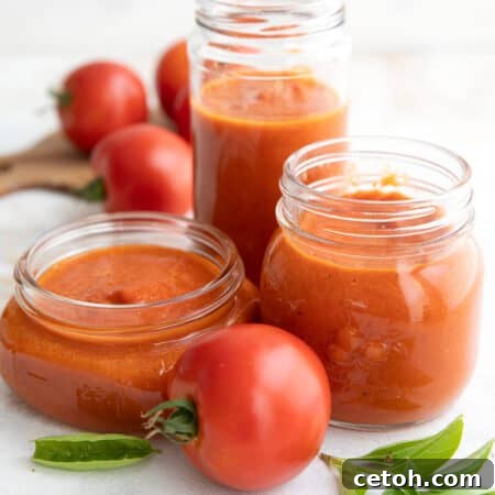 Three jars of roasted tomato sauce on a white table with fresh tomatoes and sprigs of basil.