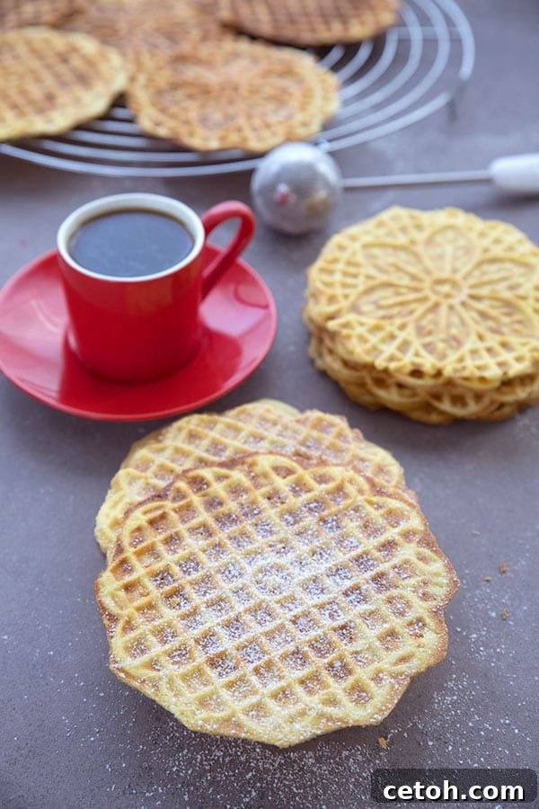 A stack of keto pizzelle cookies, lightly dusted with powdered sweetener, resting on a rustic brown wooden table beside a vibrant red cup of coffee.