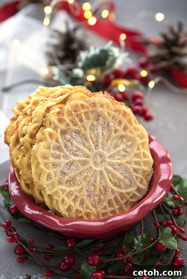 Keto pizzelle cookies arranged appealingly in a festive red bowl, adorned with sprigs of holly and pine cones, all set against a softly blurred background of warm, twinkling fairy lights.
