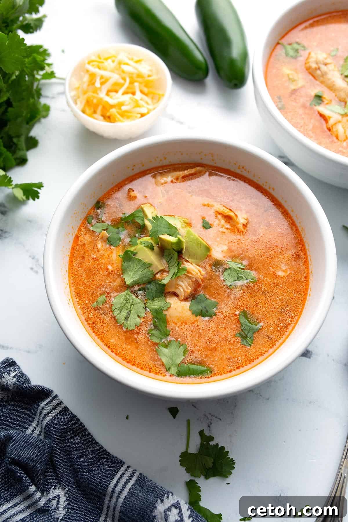 Two inviting bowls of creamy Chicken Enchilada Soup arranged on a white marble tabletop, with shredded cheese and sliced jalapeños in the background for garnish.