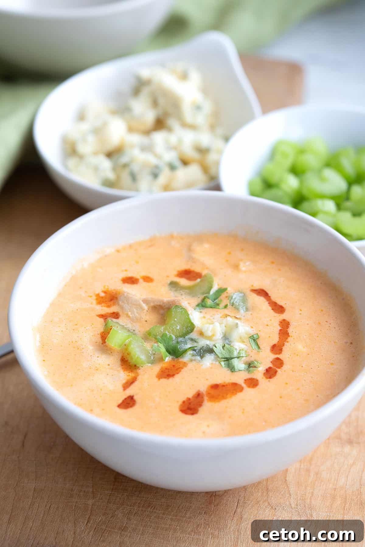 A bowl of rich Buffalo Chicken Soup sits on a rustic cutting board, with fresh celery sticks and crumbled blue cheese in the background.