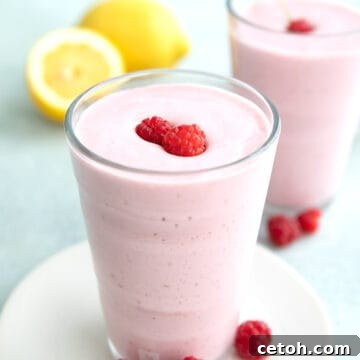 A keto protein shake in a glass on a white plate with raspberries around it and lemon in the background.