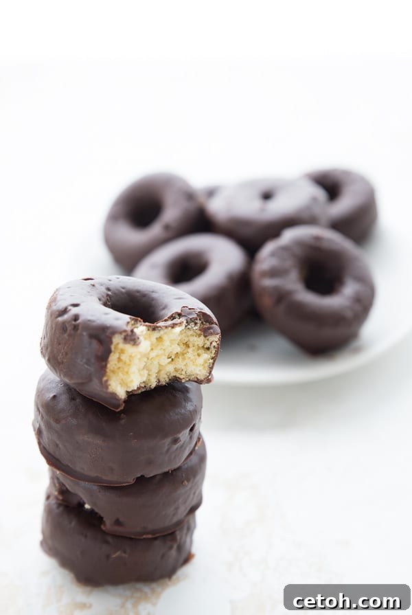 Decadent Keto Chocolate Donuts 4 A stack of keto chocolate covered mini donuts with a bite taken out of the top one. More mini donuts in the background on a white plate