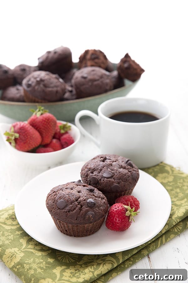 Two double chocolate zucchini muffins sit on a white plate over a green patterned napkin. A cup of coffee, a bowl of berries, and a bowl of muffins in the background.