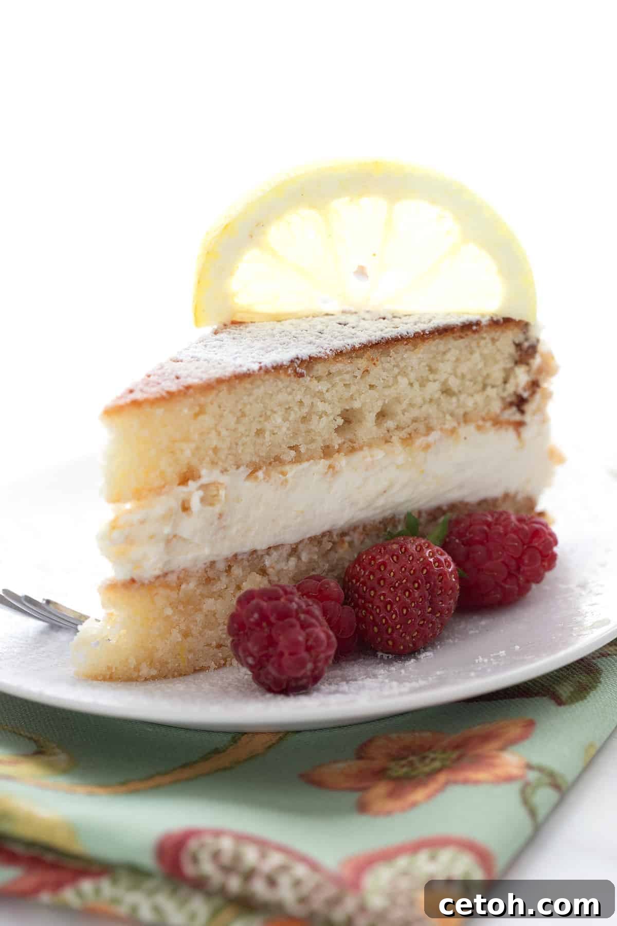 A slice of keto lemon cake on a white plate with berries on the side and a lemon slice on top.