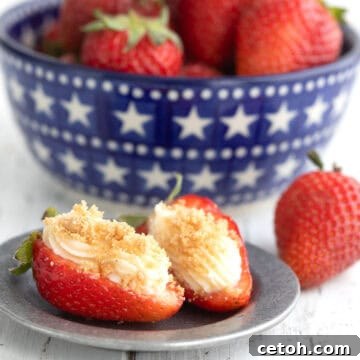 Two Cheesecake Stuffed Strawberries with graham cracker crumbs on a small pewter plate in front of a bowl of strawberries.