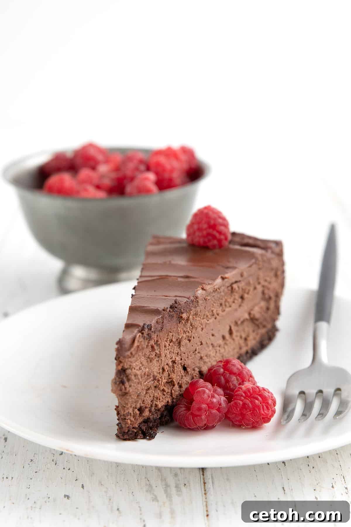 A perfect slice of Keto Chocolate Cheesecake, elegantly placed on a white plate and adorned with fresh red raspberries and a fork, ready to be enjoyed.