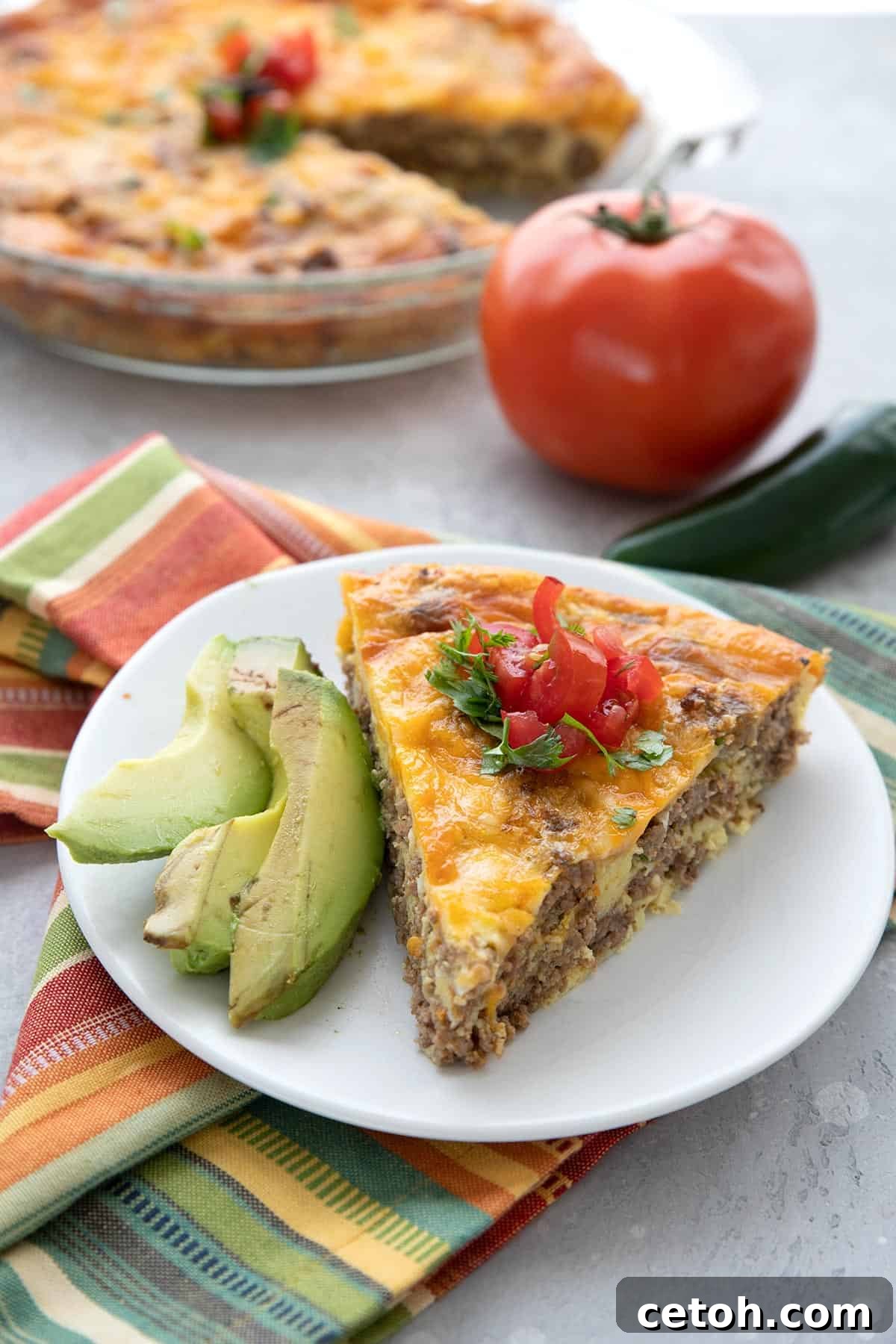 A slice of taco pie on a white plate with slices of avocado. A tomato and some jalapeno in the background.