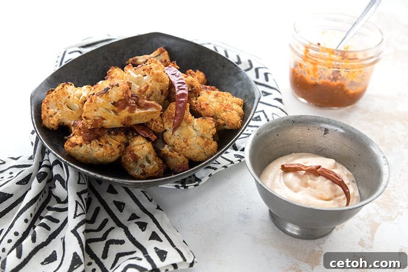 A black bowl filled with perfectly roasted air fryer cauliflower, accompanied by a jar of homemade harissa and a small bowl of creamy aioli for dipping.