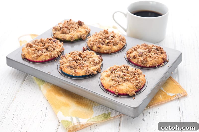 The Ultimate Keto Coffee Cake Collection 3 Close-up shot of crumb-topped keto coffee cake muffins, perfectly baked in a muffin pan with a blurred background of a coffee cup and saucer, highlighting their inviting texture.