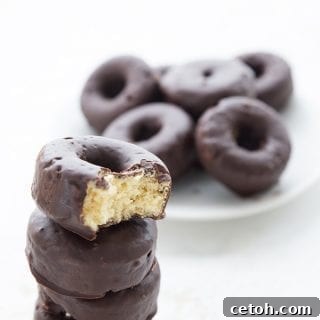 Irresistible Keto Chocolate Minis 6 A stack of keto chocolate covered mini donuts with a bite taken out of the top one. More mini donuts in the background on a white plate