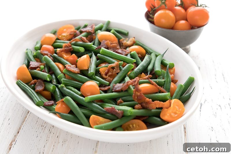 Close-up of the Keto Green Bean Salad featuring bright green beans, halved red cherry tomatoes, and crispy bacon bits, served in a white oval bowl on a clean white surface.
