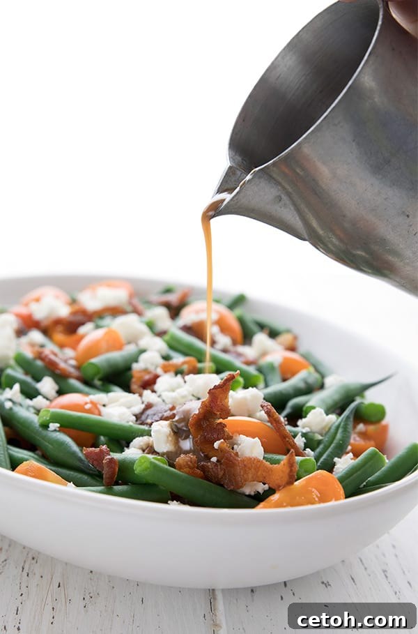 A stream of golden Dijon vinaigrette dressing being poured from a sleek pewter pitcher over a bowl of fresh green bean salad with crispy bacon.