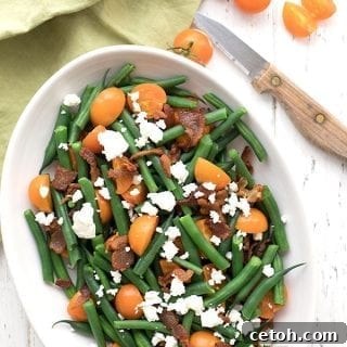 Top-down photo of the easy Keto Green Bean Salad in an oval dish on a white table, accompanied by a green napkin and some cut cherry tomatoes for garnish.
