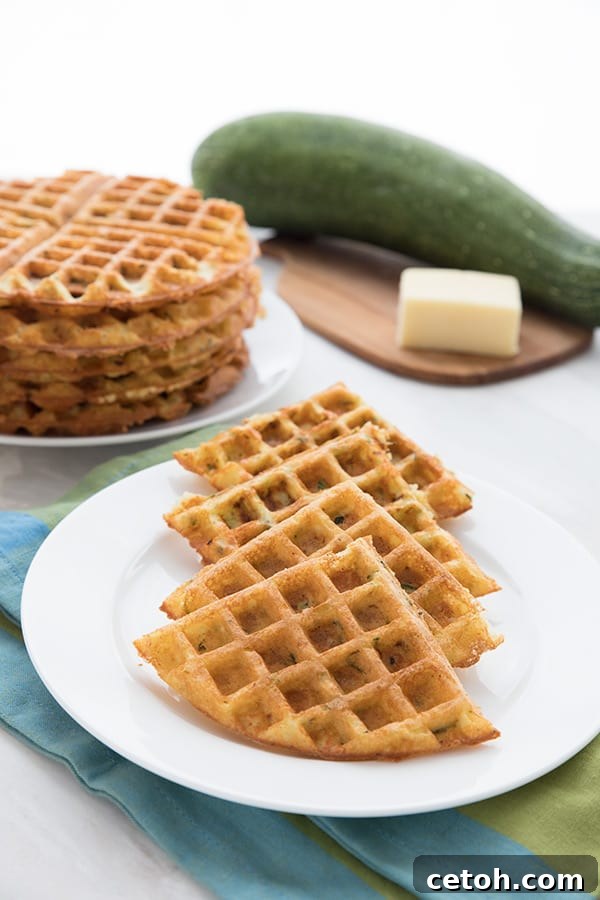A white plate filled with keto cheddar zucchini waffles. More waffles and a zucchini and some cheese on a cutting board in the background