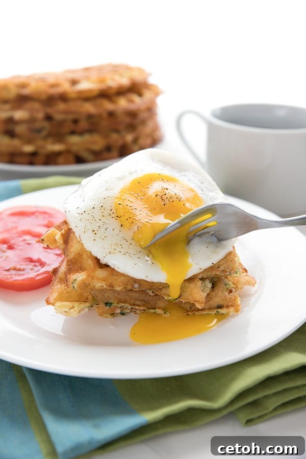 A fork going into an egg on a stack of keto cheddar zucchini waffles, allowing the yolk to run down.