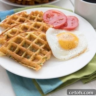 A white plate filled with eggs, zucchini waffles and tomatoes sites over a blue and green napkin