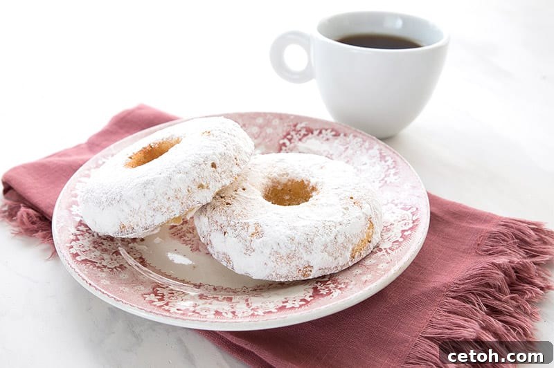Keto Couple's Kitchen 4 Delightful Keto powdered sugar donuts for two, elegantly arranged on a festive red patterned plate, resting on a complementary red napkin. A steaming cup of coffee in the background completes this inviting scene, perfect for a cozy morning treat.