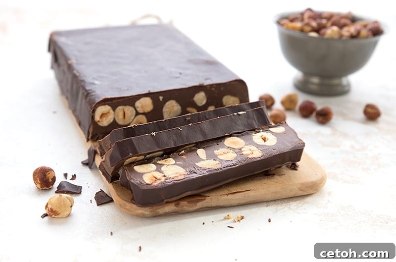 Keto chocolate hazelnut torrone cut into slices on a wooden cutting board, with a bowl of toasted hazelnuts in the background.