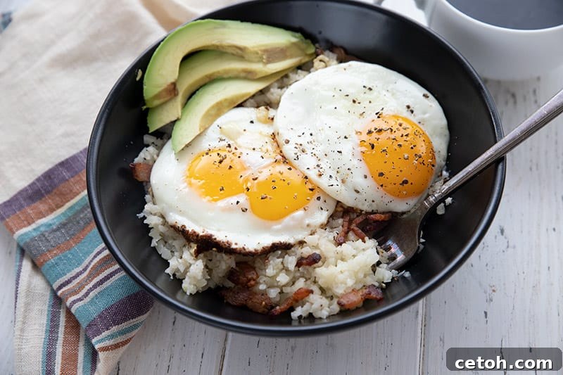Cauliflower Bacon Grits in a black bowl, topped with fried eggs and avocado