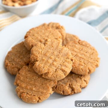 Keto Peanut Butter Cookies piled on a white plate with a small bowl of peanuts in the background.