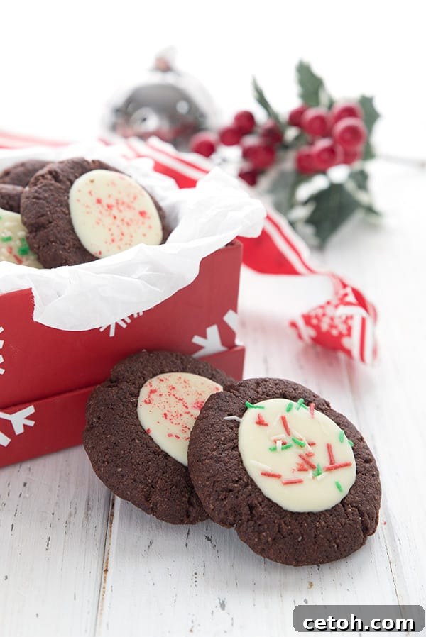 Two keto peppermint thumbprint cookies site in front of a gift box filled with more cookies and some holly in the background.