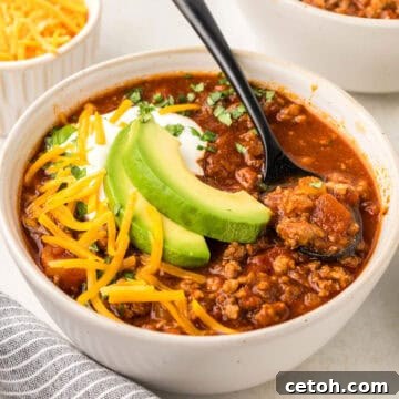 Square image of a white bowl filled with keto chili, with slices of avocado and shredded cheese on top.