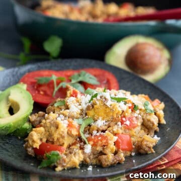 Chorizo and eggs on a black plate with avocado and tomatoes, over a striped napkin.