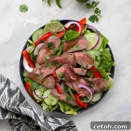 Top down image of Thai beef salad on a black plate with lime, cucumber, and herbs strewn around.
