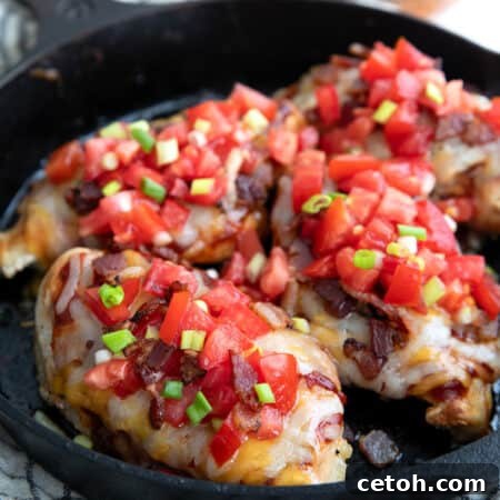 Four chicken breasts in a cast iron skillet, topped with cheese, bacon, tomatoes, and green onion.