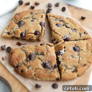 A large keto chocolate chip cookie cut into four pieces, sitting on a wooden cutting board with chocolate chips strewn around.