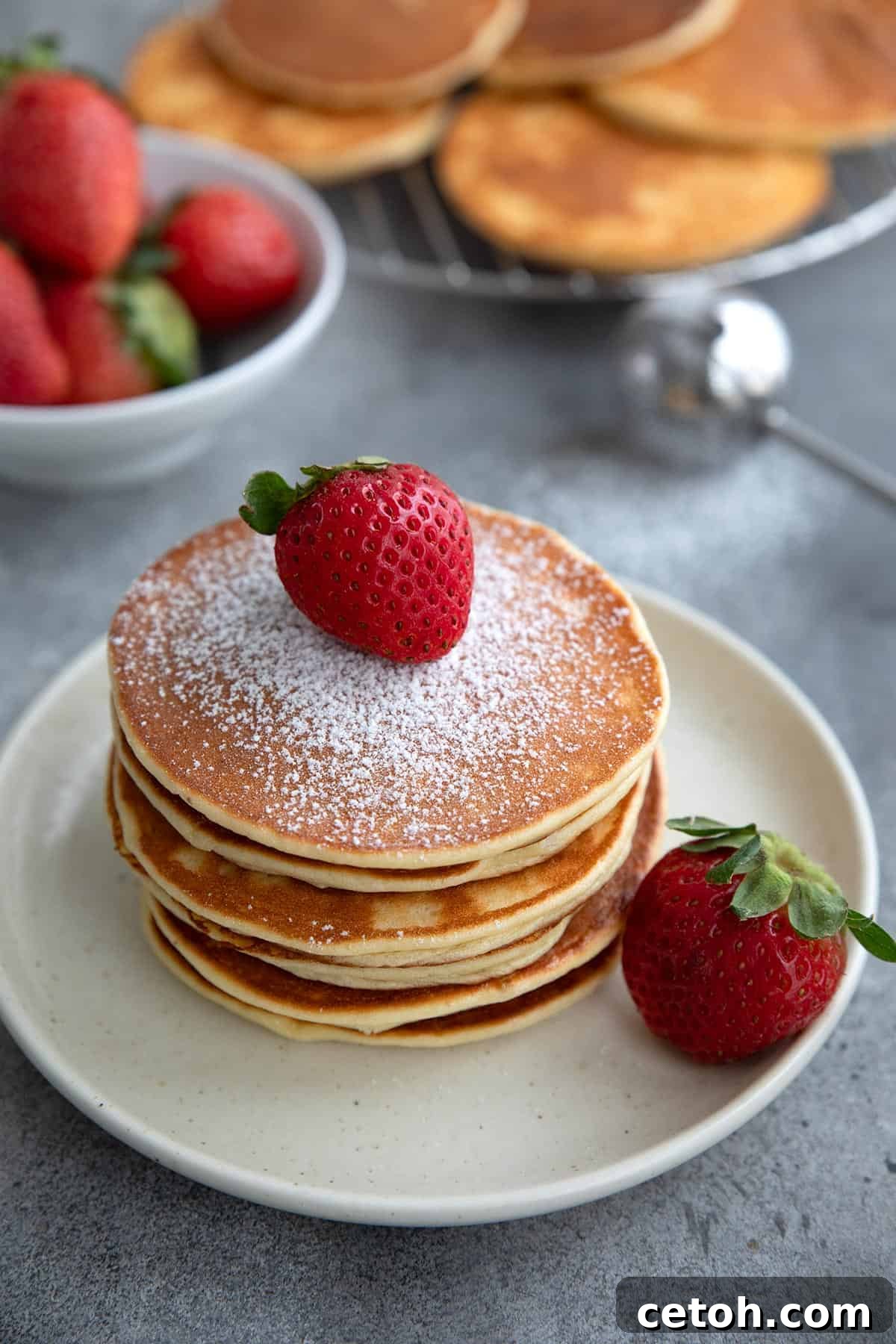 A pristine stack of keto cottage cheese pancakes elegantly displayed on a white plate, lightly dusted with powdered sweetener and crowned with a fresh, juicy strawberry.