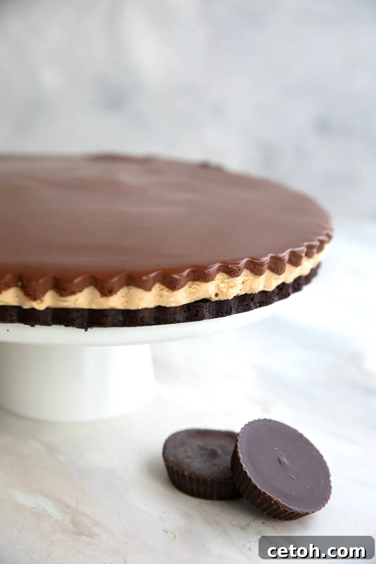 A stunning Keto Peanut Butter Brownie Tart gracefully presented on an elegant white cake stand, tastefully accompanied by two tempting peanut butter cups at its base.