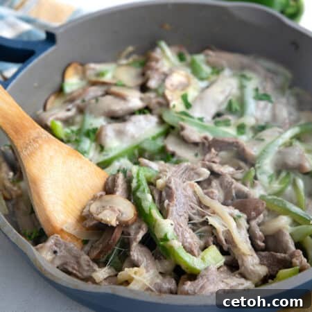 A wooden spoon digging into a pan full of Philly Cheesesteak fillings covered in melted cheese.