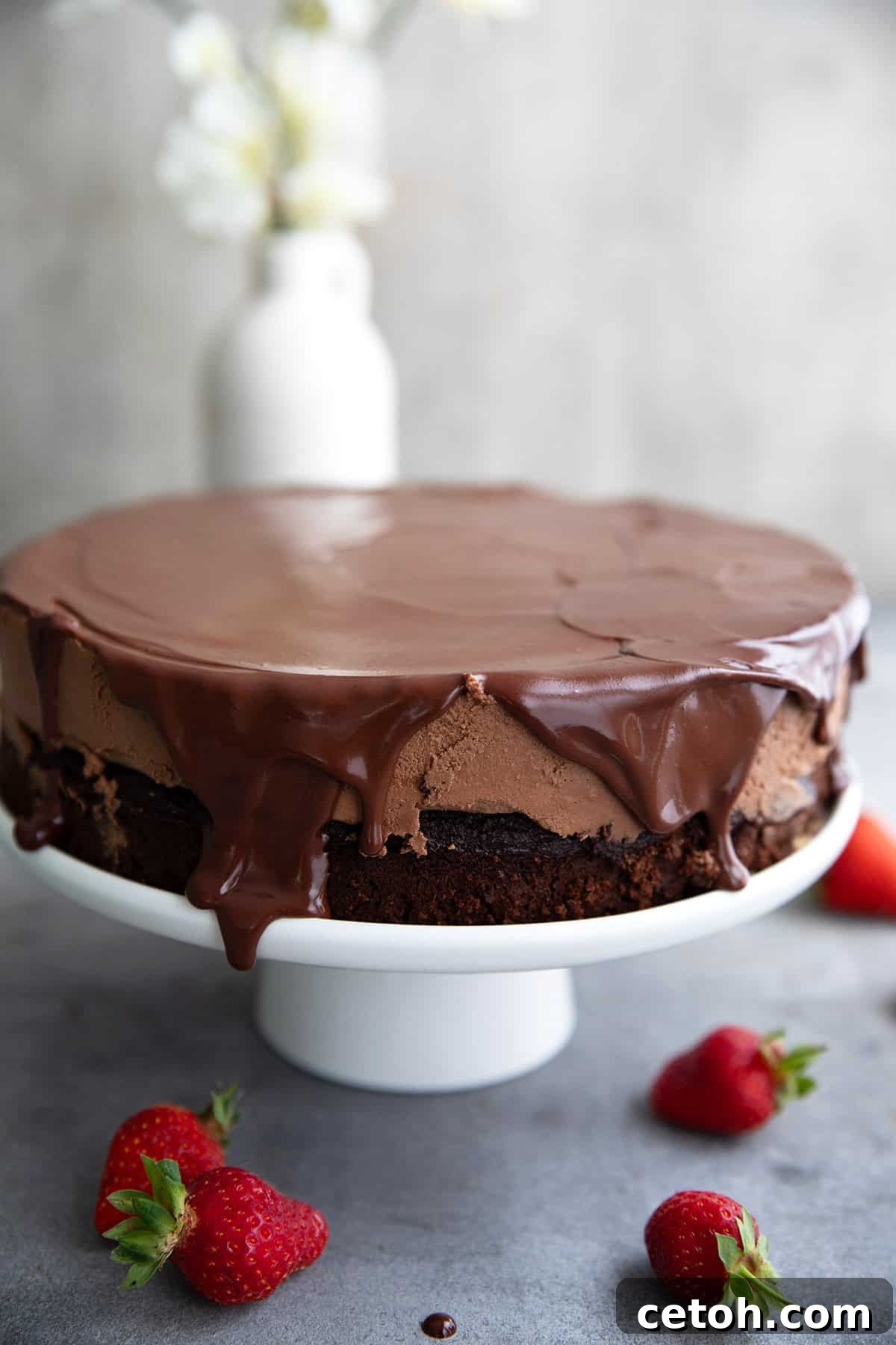 Keto Chocolate Mousse Cake on a white cake stand with chocolate glaze dripping down the cake. Strawberries are scattered around the table top.