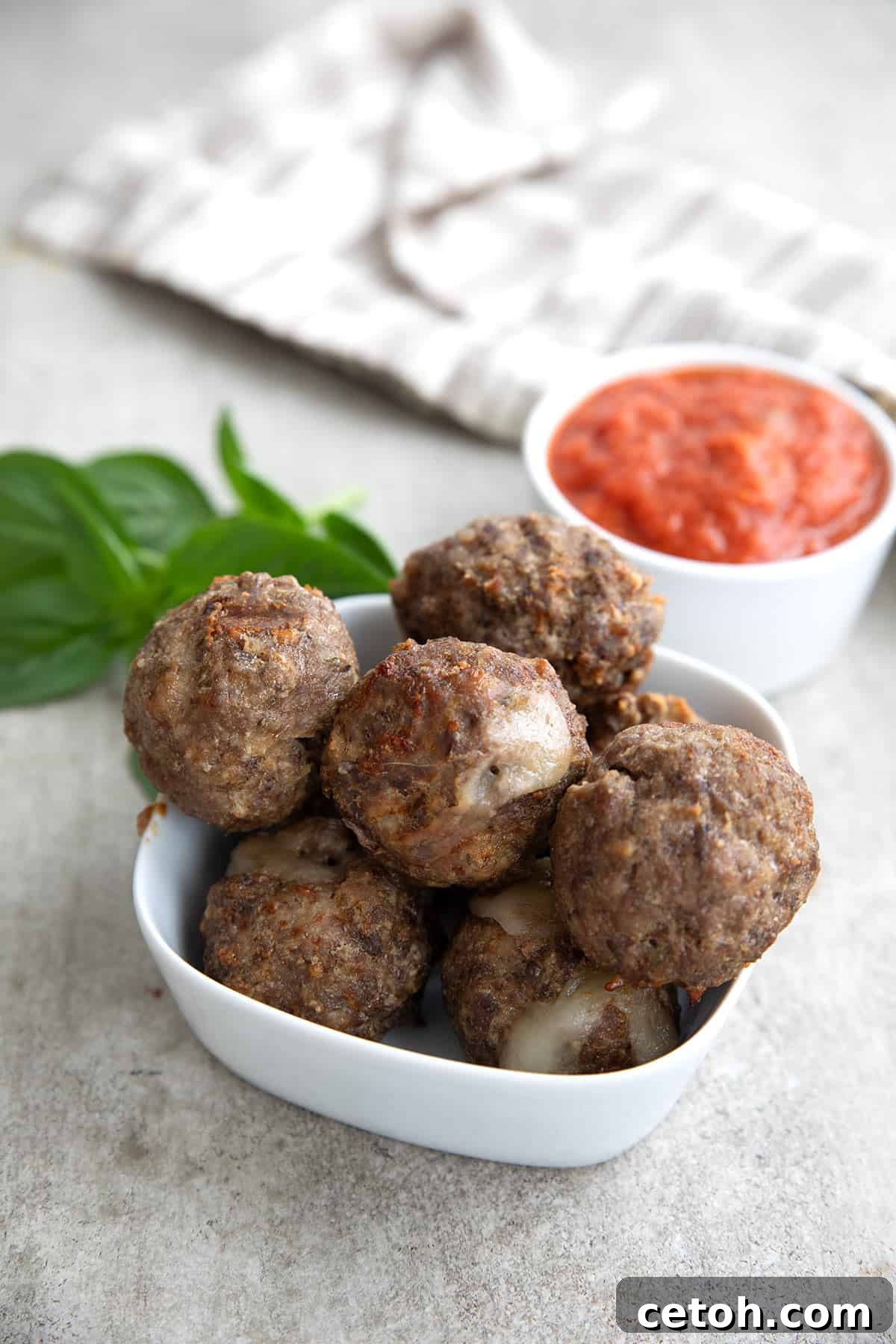 Air fryer meatballs piled up in a small white dish, with a white bowl of tomato sauce in the background.