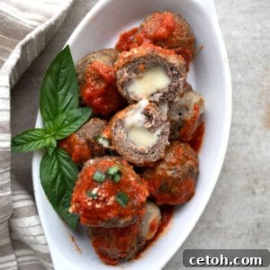 Top down image of keto stuffed meatballs with tomato sauce piled up in a white oval dish. One meatball is cut in half to show the inside.
