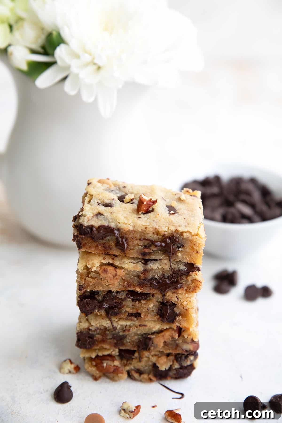 Chocolate Chip Keto Cookie Bars Loaded with Nuts 6 A stack of loaded keto cookie bars on a white table with a bowl of chocolate chips and a vase of white flowers in behind.